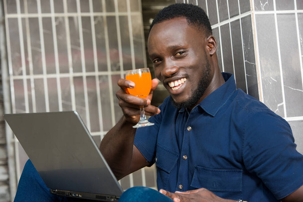 young man in blue shirt sitting near a wall looking at laptop smiling with a glass of juice in hand. - Fotó, kép