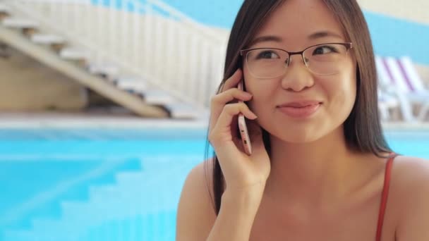 Enfocado frunciendo el ceño asiático millennial empresaria habla por teléfono inteligente, resuelve problemas de negocio sentado en la piscina en el complejo de spa o se comunica con amigos con tecnología de red móvil, primer plano - Metraje, vídeo