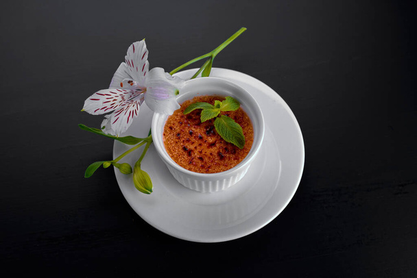 Dessert creme brulee, in a white dish with mint leaves in white flowers, on a dark background, with a place for text - Photo, Image