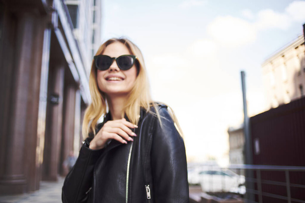 Fashionable woman in a leather jacket and sunglasses near the building on the street - 写真・画像