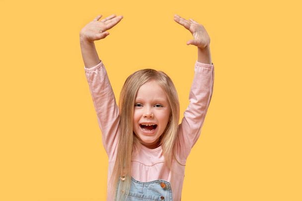 Cheerful beautiful blonde girl happily raised her hands up. Yellow background. - Фото, изображение
