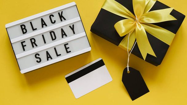 Banner concept for Black Friday celebration,lettering.White board with text BLACK FRIDAY SALE,black gift box with yellow bow,tag,credit card on yellow background,top view,flat lay,close-up - Photo, Image