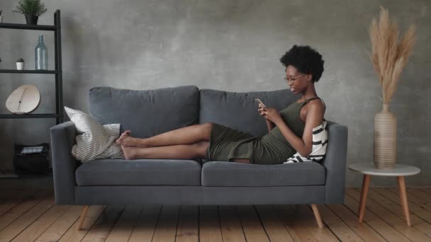 Gelukkig Afrikaans Amerikaanse jonge vrouw zitten ontspannen op gezellige bank blij om te verhuizen naar een nieuw appartement. Glimlachend zwart meisje rust op comfortabele bank in de woonkamer dromen - Video