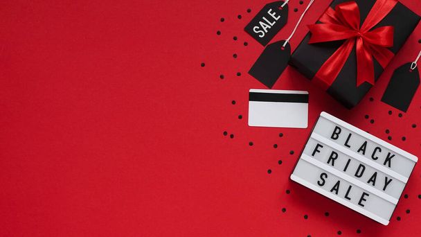 Banner concept for Black Friday celebration, lettering.White board with text BLACK FRIDAY SALE, gift box with red bow, tag, credit card, confetti on red background, top view, flat lay, close-up, copy space. - Фото, изображение