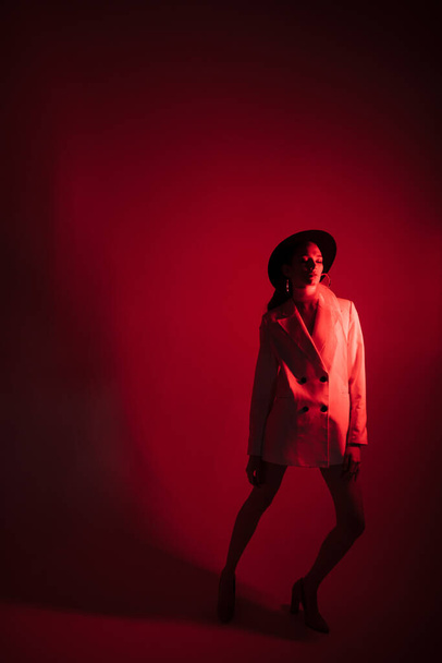 Young girl in a white suit and a black hat. The concept of clothing. Red light. Red background - 写真・画像