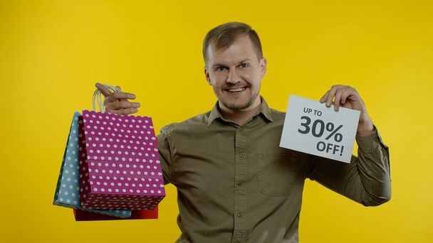 Mężczyzna pokazuje się do 30 procent off napis i torby na zakupy, wygląda zadowolony z niskich cen - Zdjęcie, obraz