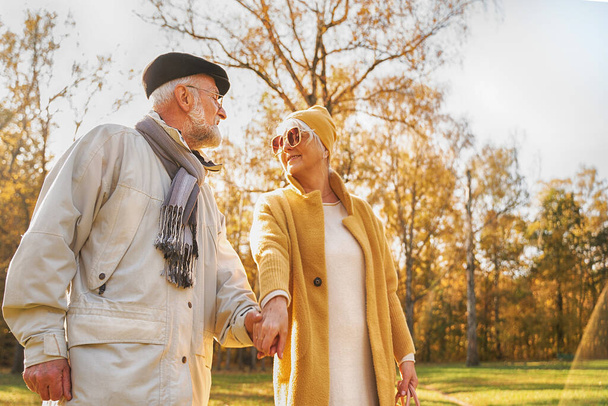 Romantic senior couple walking in beautiful autumn city park, holding hands. Elderly woman looking at her husband with love and smile. Relationship and people concept  - Фото, изображение