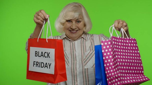 Elderly grandmother in shirt showing Black Friday inscription on shopping bags, rejoicing discounts - Foto, imagen