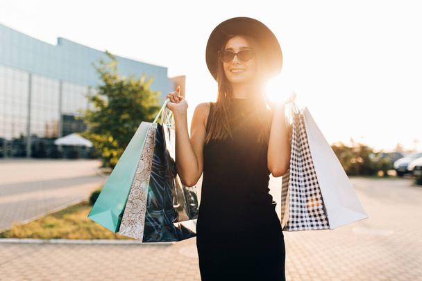attractive elegant girl, in a black dress and hat, wearing dark glasses, holding shopping bags and enjoying shopping, stands in the city against the background of the sunset. Black Friday, shopping - 写真・画像