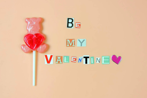 Be my Valentine - words made of paper cut letters and lollipop bear with heart on pink background - Photo, image