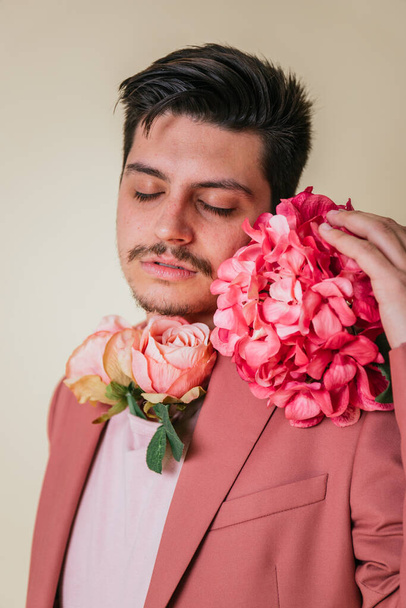 Portret przystojnego chłopca z zamkniętymi oczami. Przystojny młody mężczyzna z kwiatami na szyi i obok twarzy w studiu fotograficznym na żółtym tle w różowym garniturze i koszulce. - Zdjęcie, obraz