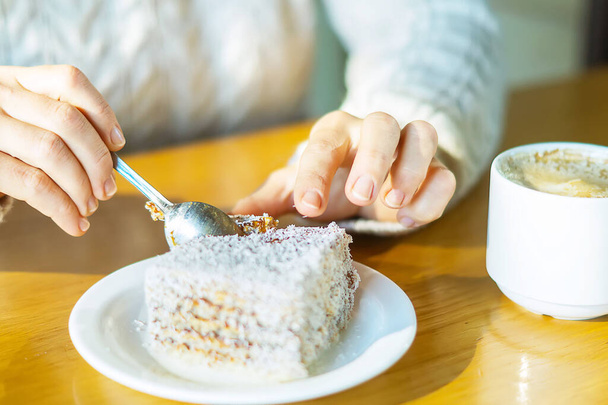 Women's hands in a warm knitted sweater in a city cafe with a teaspoon cut off a piece of delicious cake that lies on a white saucer. Coffee mug on the table. Relax in the cafe. selective focus. - Foto, Imagem