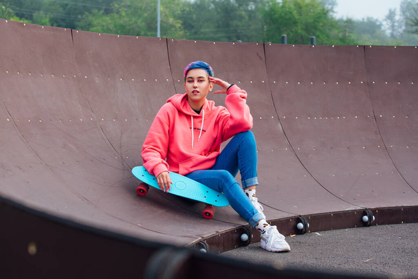 Stylish young woman with short colored hair sitting with her blue plastic skateboard in skatepark. Youth concept. - Φωτογραφία, εικόνα