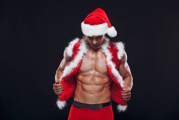 Sexy Santa Claus. Bodybuilder young handsome santa clause smile wearing Santa Claus hat shows off abs cubes at New Years eve and Christmas winter holiday black background. - Fotografie, Obrázek