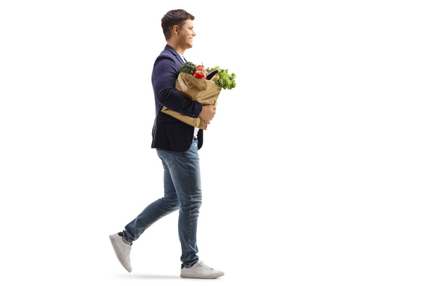 Full length profile shot of a smiling young man walking and carrying a paper bag with groceries isolated on white background - Φωτογραφία, εικόνα
