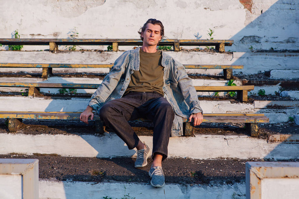 Fashion portrait of a tall handsome man dressed in jeans jacket on the crushed tribune of the old stadium during sunset - Foto, Imagen