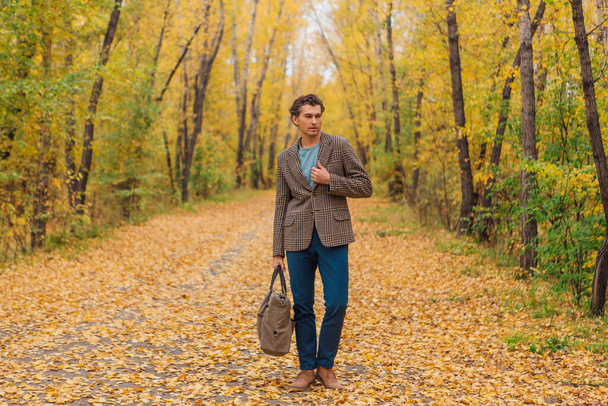 Tall handsome man dressed in a brown jacket walking with a bag on the autumn alley - Photo, image