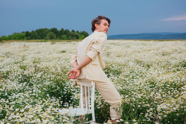 Tall handsome man dressed in a white suit on naked body sitting on a back of a chair in camomile flowers field - Photo, image