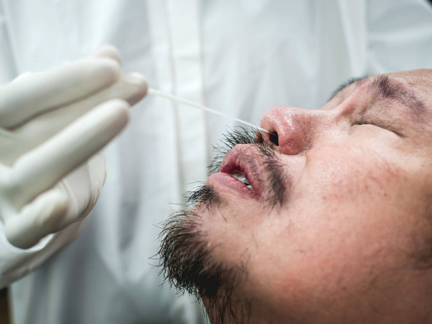 アジア人男性は、彼の鼻に部分的に挿入された軍不快な鼻咽頭スワブ迅速なテストを受けます。本物のcovid鼻波検査と患者からの反応。白衣を着た匿名の看護師. - 写真・画像