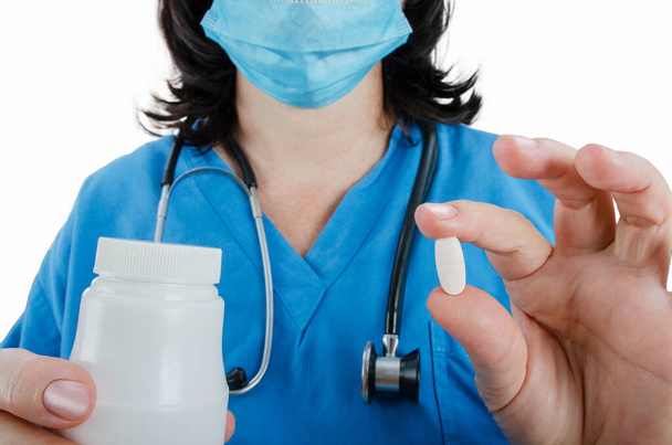 Το χέρι ενός γιατρού με μπλε στολή κρατάει ένα λευκό χάπι. Από την άλλη πλευρά, είναι ένα λευκό πλαστικό βάζο. Κοντινό πλάνο, ακέφαλο. - Φωτογραφία, εικόνα
