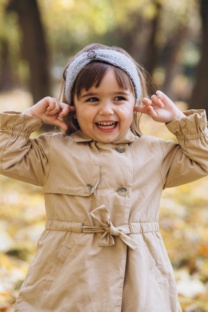 Onnellinen ja kaunis tyttö beige takki näyttää tunteita ja sulkee korvansa sormillaan syksyllä puistossa. - Valokuva, kuva