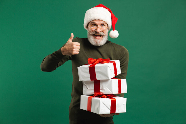 Bearded modern Santa Claus holding gifts and smiling on the green background - Photo, Image