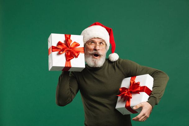 Der bärtige moderne Weihnachtsmann hält eine Geschenkschachtel in der Hand und lauscht auf dem grünen Hintergrund, was drin ist - Foto, Bild