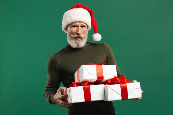 Vousatý moderní Santa Claus drží dárky a usmívá se na zeleném pozadí - Fotografie, Obrázek