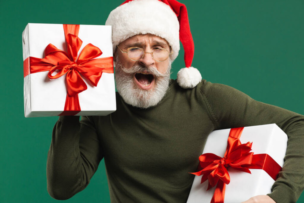 Bearded modern Santa Claus holds a gift box and listens to what's in it on the green background - Fotoğraf, Görsel