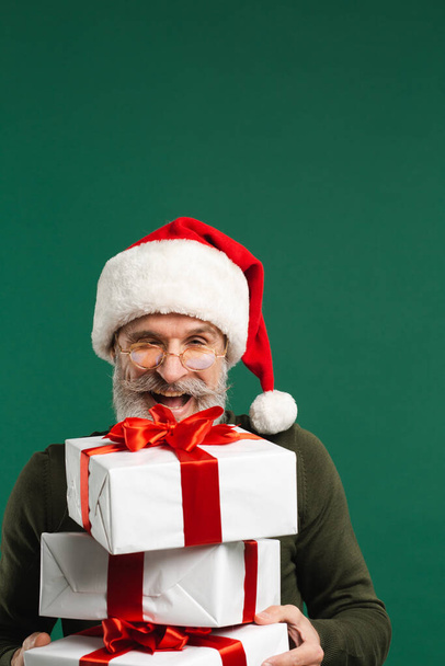 Brodaty nowoczesny Święty Mikołaj trzyma prezenty i uśmiecha się na zielonym tle - Zdjęcie, obraz