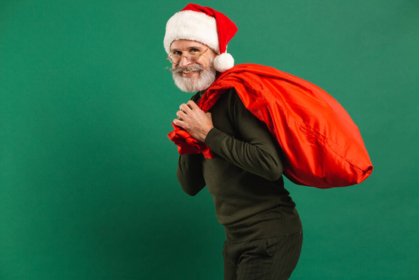 Happy bearded modern Santa Claus holding red bag on the green background - Photo, Image