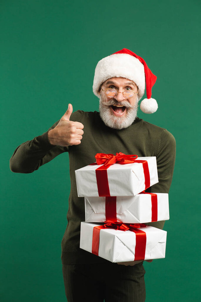 Brodaty nowoczesny Święty Mikołaj trzyma prezenty i uśmiecha się na zielonym tle - Zdjęcie, obraz