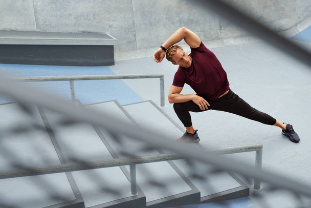 Young Man Stretching Near Concrete Steps Outdoors. Handsome Caucasian Sportsman With Strong Muscular Body In Fashion Sportswear Warming Up Before Intense Workout. - Фото, изображение