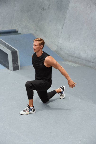 Young Man Warming Up Against Concrete Wall Outdoors. Handsome Caucasian Sportsman With Strong Muscular Body In Fashion Sportswear Exercising Before Intense Workout. - Photo, Image