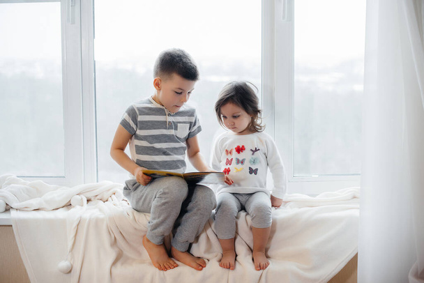 Brother and sister are sitting on the windowsill and reading a book. Happiness, family - Photo, image
