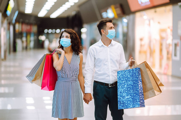 Beautiful couple in protective medical mask shopping in mall. Happy  couple holding shopping bags, looking upon showcase and smiling while standing in mall. Covid-2019. - Φωτογραφία, εικόνα