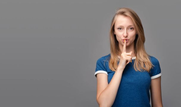Pretty young blonde woman, dressed in blue t shirt, keeps finger on lips, making hush gesture on gray background. Shh, silence concept. Copy space for your text - Foto, Imagem