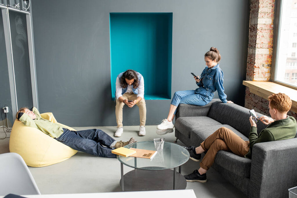 Három fiatal pihentető tervezők vagy fejlesztők mobil alkalmazások gördülő okostelefonok, míg a szőke nő feküdt sárga karosszék - Fotó, kép