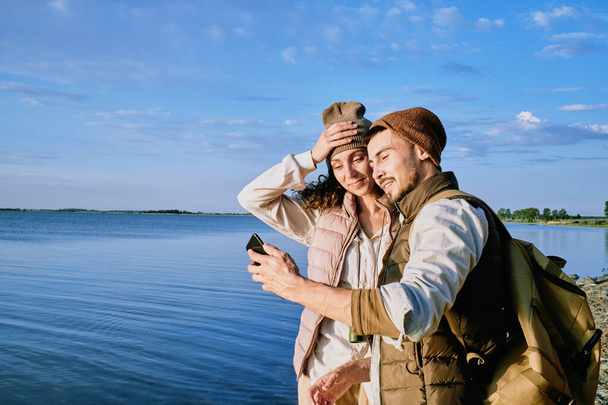 Contenido hermosa pareja joven en sombreros y chalecos de pie en la orilla del lago y tomar selfie en el teléfono inteligente durante la caminata - Foto, imagen