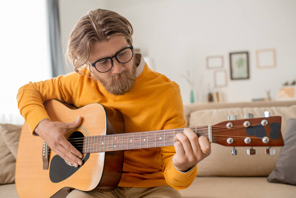Современный молодой человек в джинсах и желтом свитере сидит на диване и дергает за струны гитары перед камерой смартфона дома - Фото, изображение