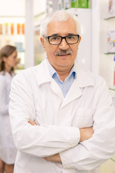 Trabajador de farmacia envejecido en blanco y anteojos cruzando brazos por el pecho mientras está de pie contra las pantallas con productos médicos y su colega - Foto, Imagen