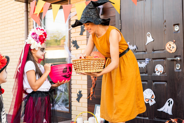 Young cheerful woman in yellow dress and black witch hat bending by cute halloween girl with basket and giving her some sweets and cookies - Photo, Image