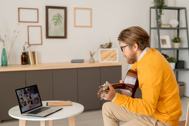 Exhibición del ordenador portátil con la muchacha adolescente diligente que juega la guitarra mientras que se sienta en el sofá en el ambiente casero durante la lección en línea con el profesor - Foto, Imagen