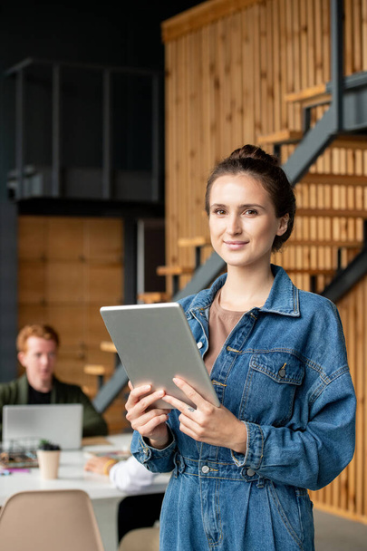 Mladá úspěšná podnikatelka s tmavými vlasy stojící před kamerou v pracovním prostředí a pomocí touchpad v kanceláři - Fotografie, Obrázek