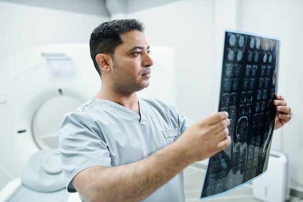 Médico pensativo y enfermera en máscaras quirúrgicas analizando la imagen de rayos X mientras el paciente duerme en la habitación del hospital - Foto, Imagen