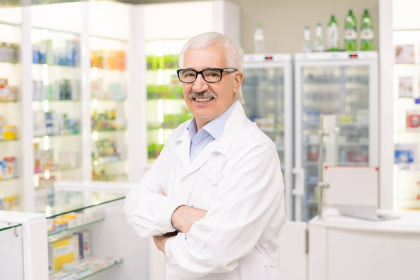 Ikääntynyt apteekin työntekijä valkoisessa takissa ja silmälaseissa, jotka ylittävät käsivarret rinnassa, kun katsot sinua hymyillen lääketieteellisten tuotteiden näyttöjä vastaan - Valokuva, kuva