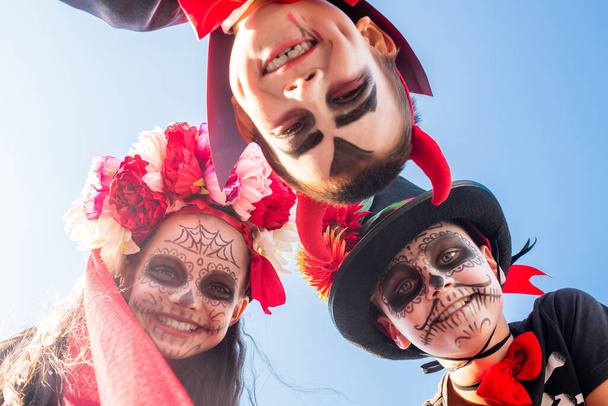 Drei glückliche Halloween-Kinder mit aufgemalten Gesichtern, die vor der Kamera vor blauem Himmel stehen und einen mit einem verschmitzten Lächeln anschauen - Foto, Bild