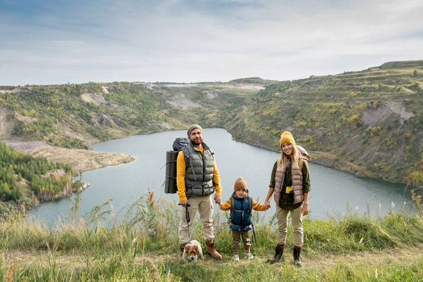 Szczęśliwa rodzina młodej pary i ich synka w ciepłym stroju casualwear stojących na chodniku przed jeziorem i górami podczas podróży - Zdjęcie, obraz