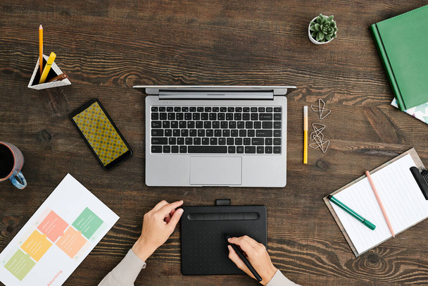 Обзор рук современного креативного дизайнера, сидящего за деревянным столом перед ноутбуком и использующего стилус и графический планшет - Фото, изображение