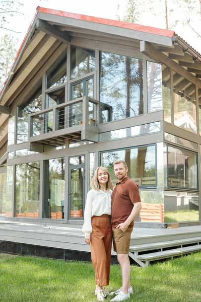Ritratto di giovane coppia sorridente in abiti casual in piedi contro la casa di vetro e abbracciarsi - Foto, immagini